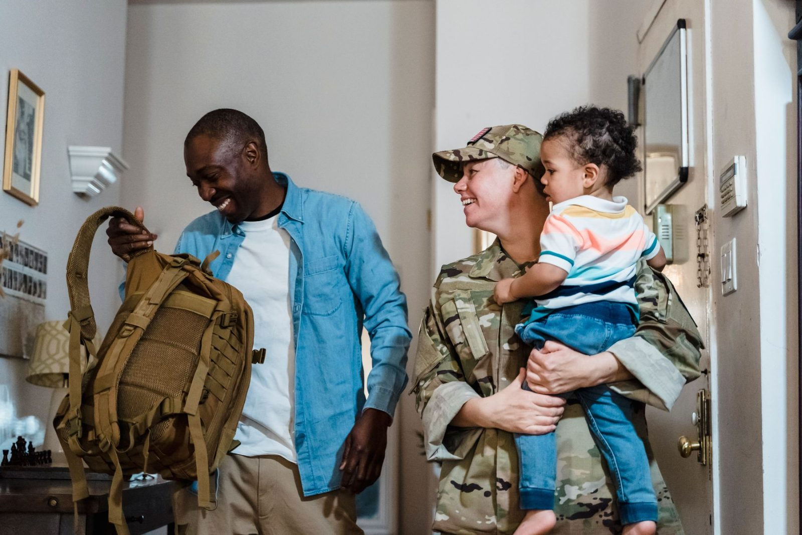 Service member in uniform holding a toddler beside her smiling spouse inside their home